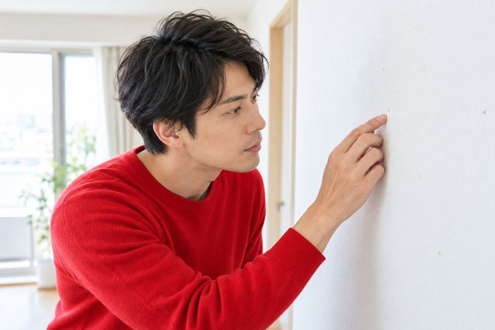 30代の男性が壁の前に立ち、ポスターを貼った跡や小さな穴がないかを近くで見て確かめている