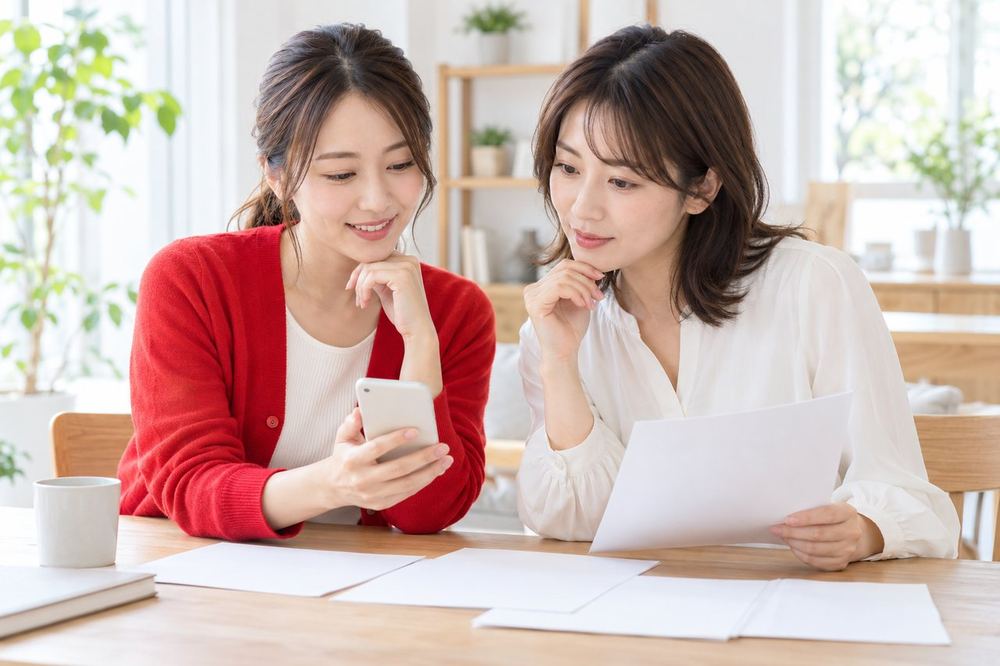 明るい部屋で、女性ふたりが机で書類とスマホを見ながら、お金の手続きについて落ち着いて話している