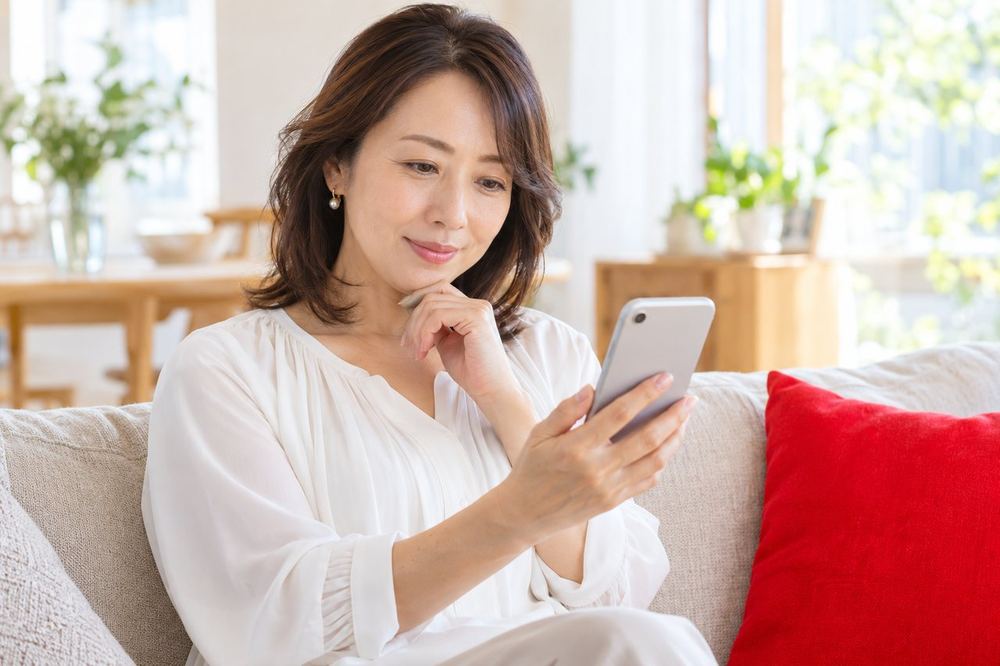 40代の女性が自宅のソファでスマホのLIVE画面を見ながら、配信者の話し方に注目している