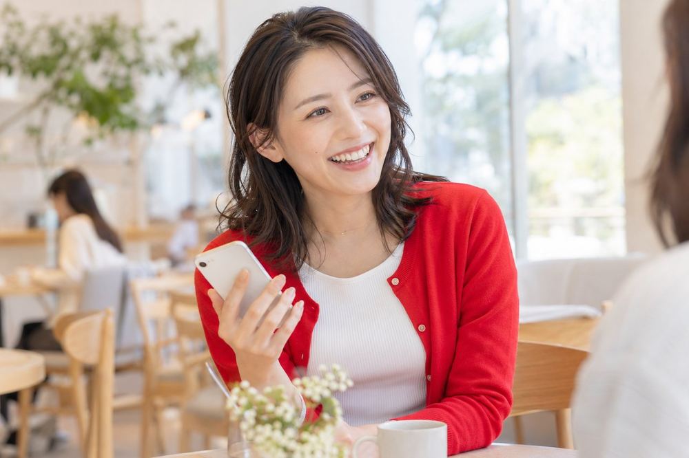 30代の女性がカフェの席でスマホを手に、笑顔で誰かと話している