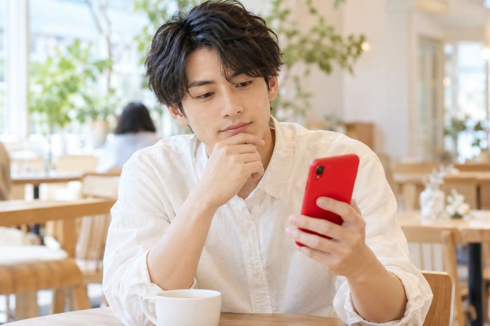 男性がカフェでスマホを見ながら、配信で顔を出すかどうか考えている