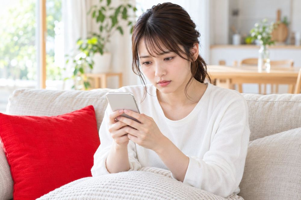 20代の女性が部屋のソファでスマホを胸の前に持ち、画面を見ながら少し不安そうに考え込んでいる
