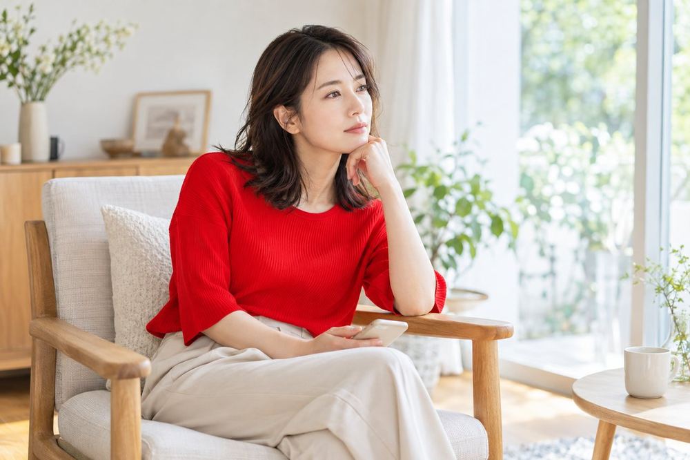 30代の女性が部屋の椅子に座り、スマホを膝に置いたまま少し考え込んでいる