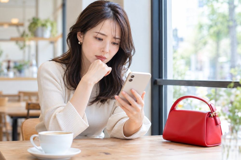 女性がカフェの窓際で、スマホを見ながら少し不安そうに考えこんでいる