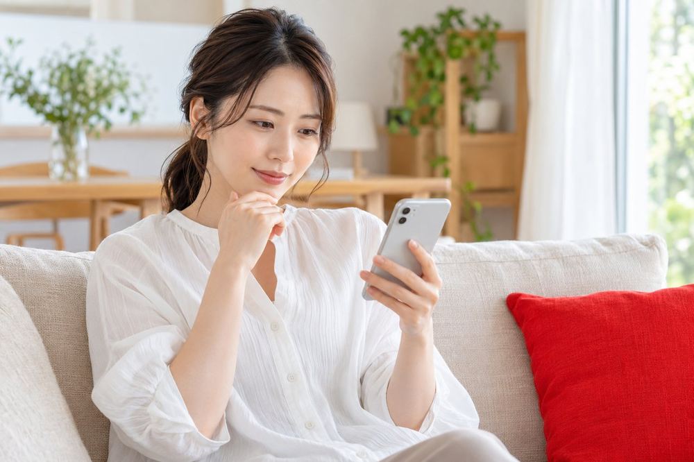 女性が自宅のソファで、友人からのメッセージを読みながら、落ち着いて返信の文面を考えている