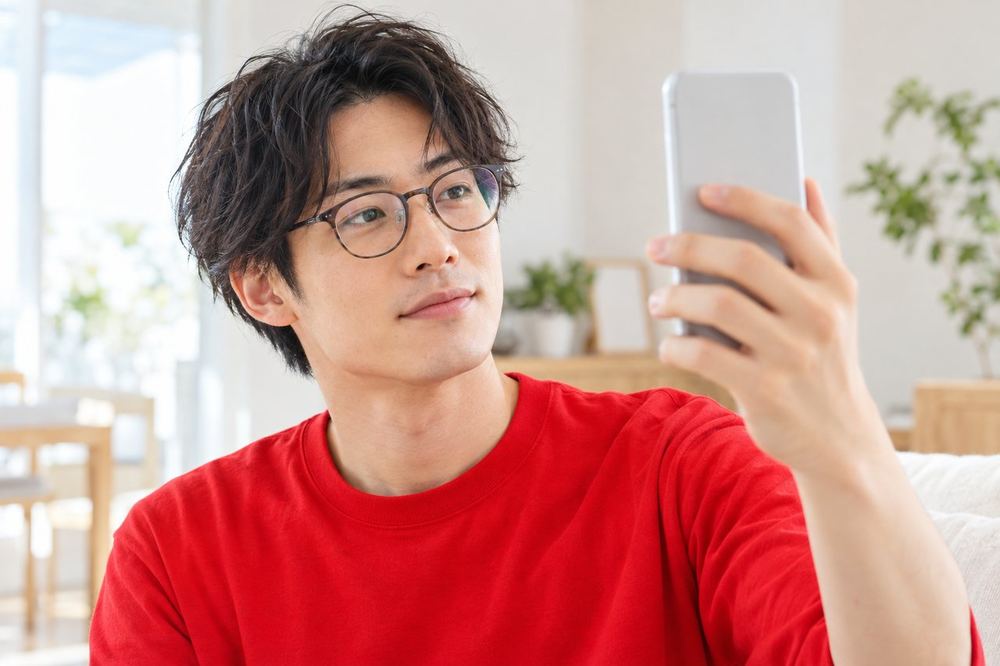 30代の男性がメガネをかけたままスマホのカメラに顔を向け、レンズに反射する光を確かめている