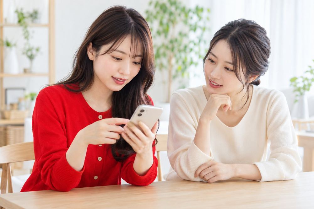 20代の女性が机でスマホを見ながら、落ち着いた表情で誰かと相談している