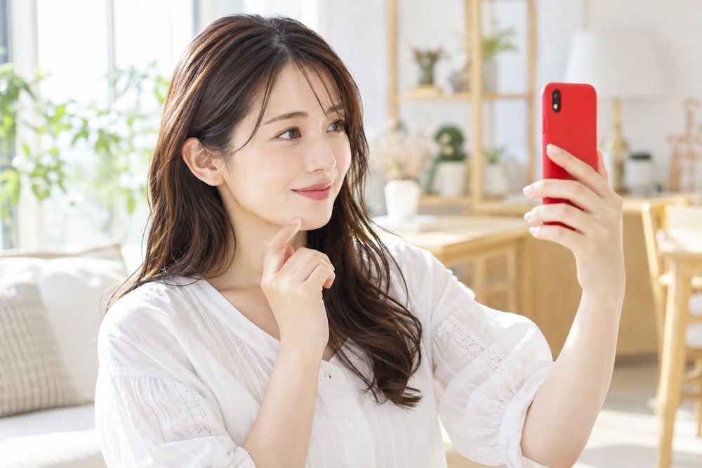 明るい部屋で女性がスマホのカメラに映る自分の顔を見ながら、口元と目元の動きを確かめている