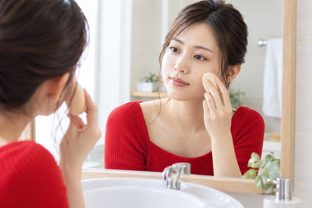 20代の女性が洗面台の鏡の前で、スポンジを使って頬にファンデーションを薄くのばしている