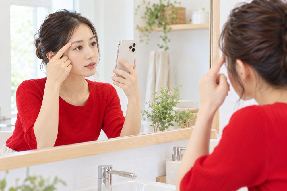 女性が洗面台の鏡の前で、眉の形を確かめながらスマホのカメラにも映して比べている