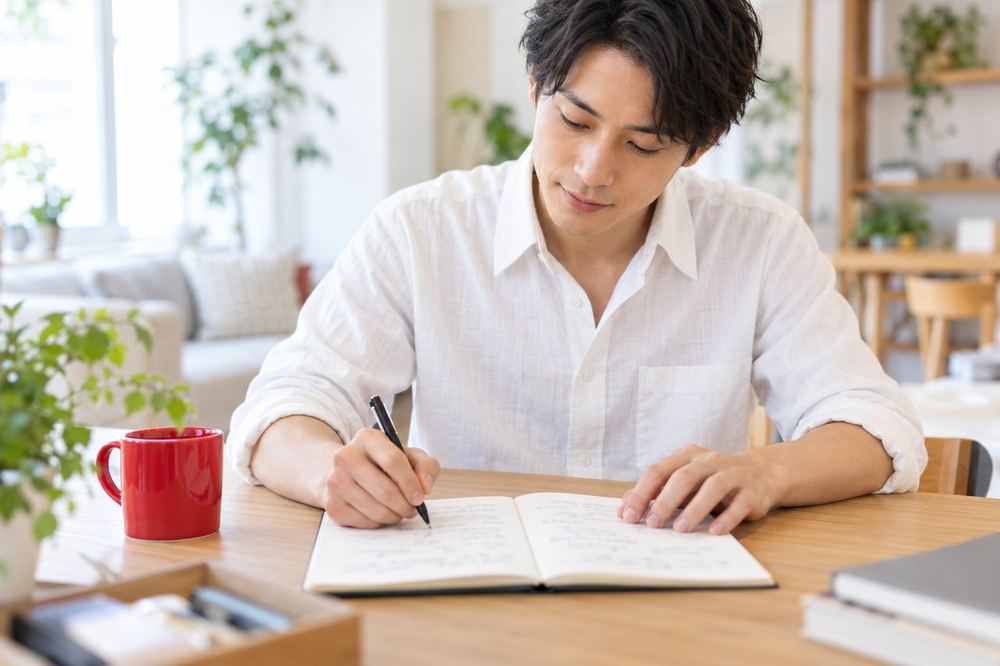 30代の男性が机にノートを広げ、バイトと配信の違いを二つの列に分けて書き出している