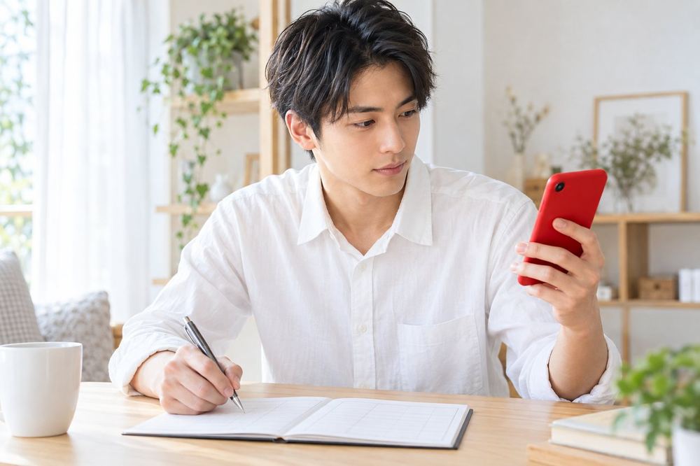 20代の男性が机でスマホの入金履歴を見ながら、月ごとの収入と経費の表に数字を書き込んでいる