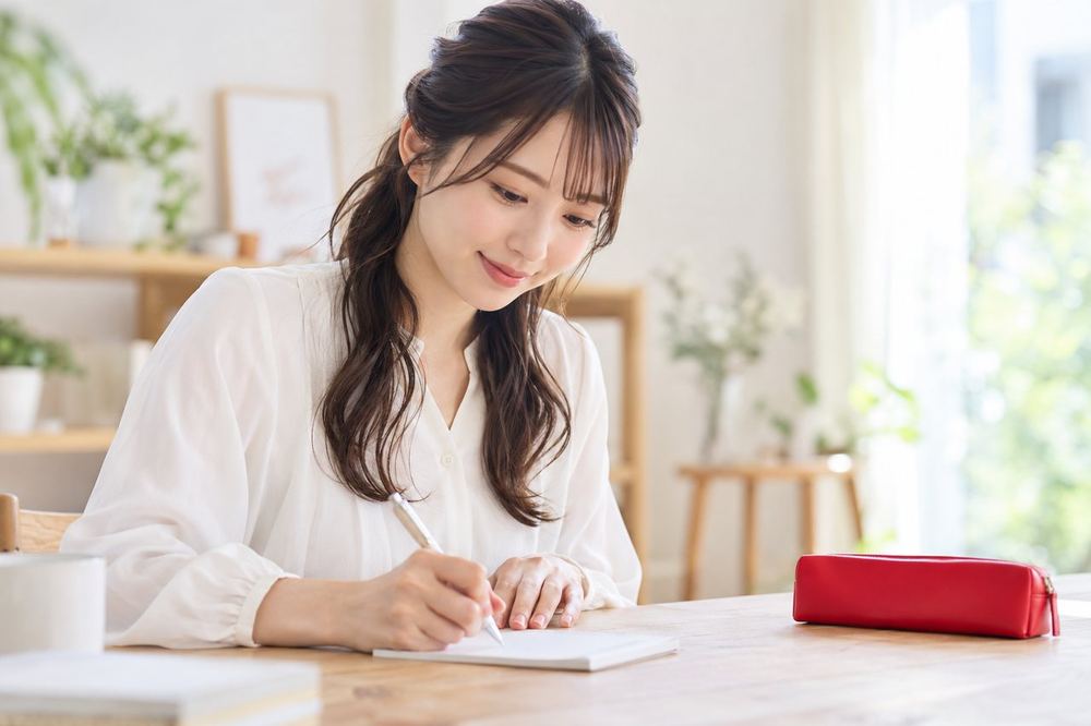 明るい部屋の机で、20代の女性がメモ用紙に、自己紹介の一言と今日の話題を箇条書きで書き出している
