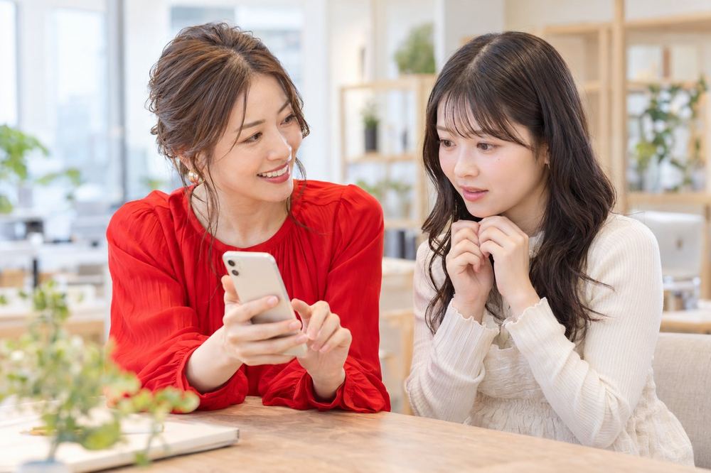 明るいオフィスで、ライバー経験のある女性のマネージャーが、緊張した様子の20代の女性に、笑顔でスマホの画面を見せながら話している