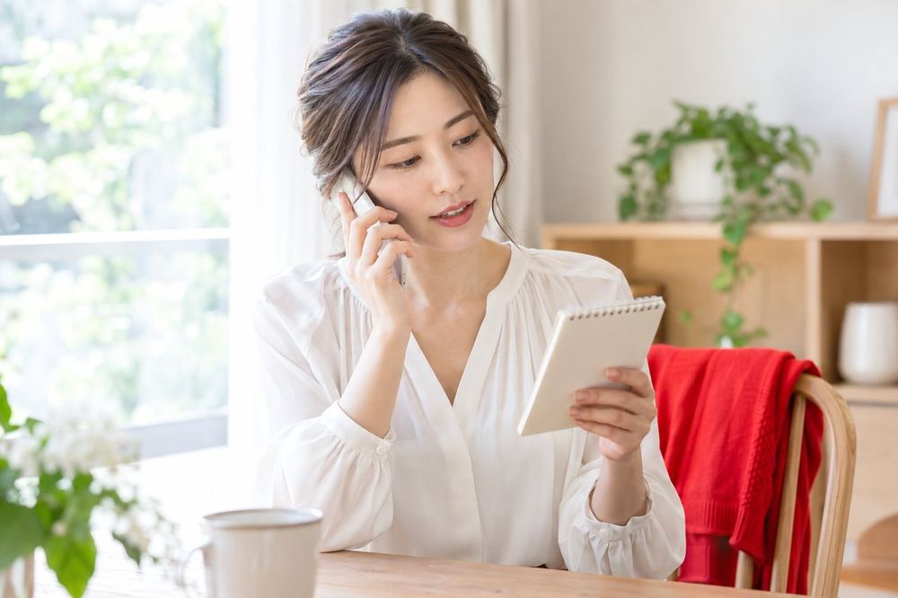 女性が窓際で電話をかけながら、メモを見て状況を整理して話している
