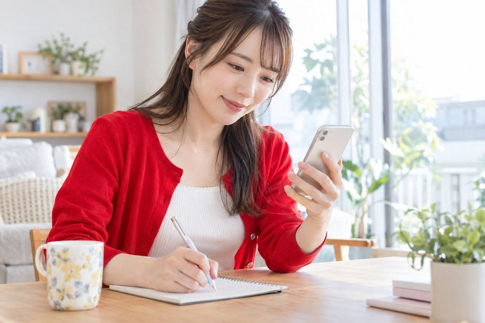 窓際の席で、女性がスマホのLIVE報酬の画面を見ながら、ノートに数字を書き留めている