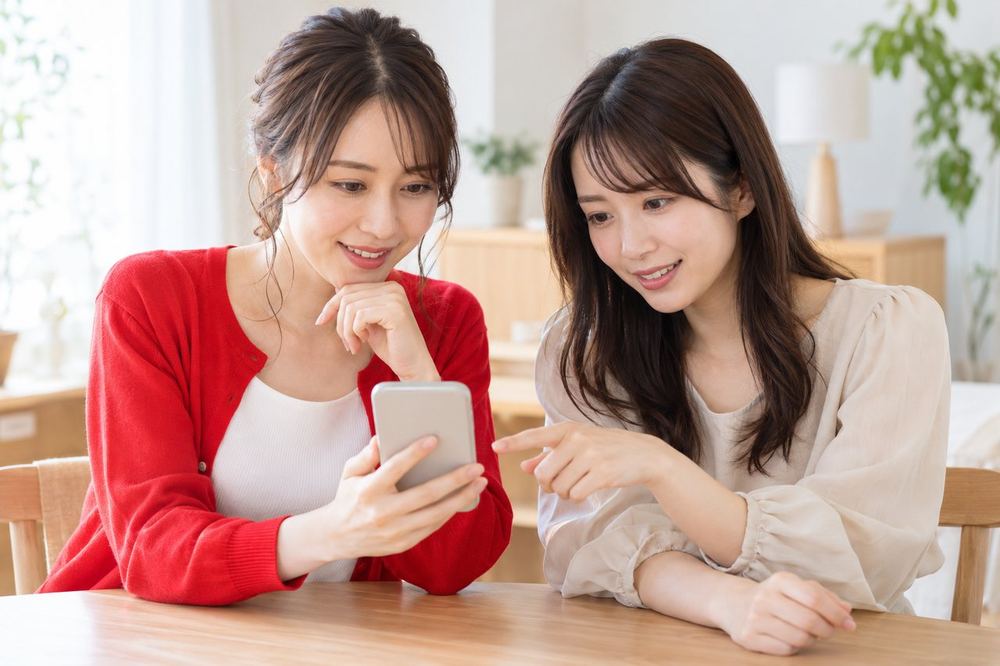 明るい部屋で、女性ふたりがスマホを見ながら、事務所の面談で聞くことを相談している