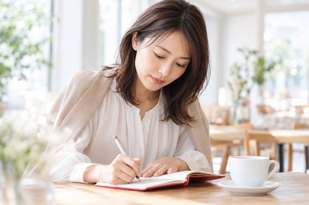 30代の女性がカフェの席で、事務所の担当者との面談に持っていく質問をノートに書き出している