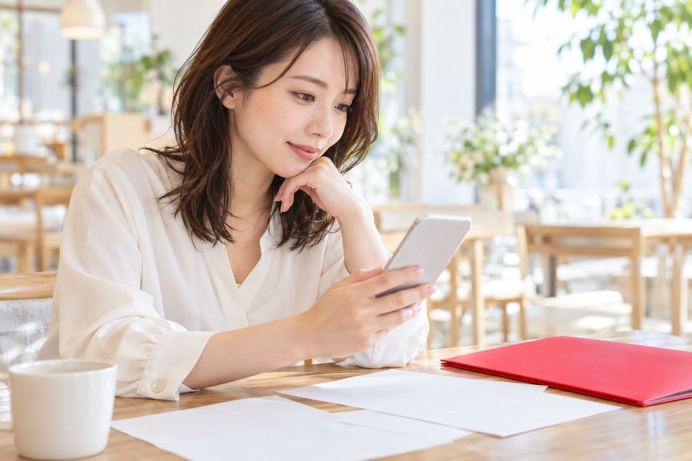 女性がカフェのテーブルで、スマホの画面を見ながら資料を広げて事務所のことを調べている