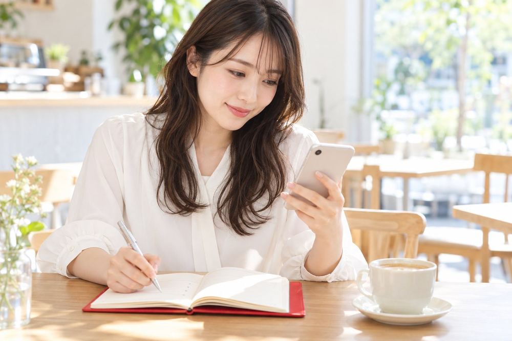 20代の女性がカフェの席で、ノートに「相談したいこと」を箇条書きにしながらスマホで連絡を取っている