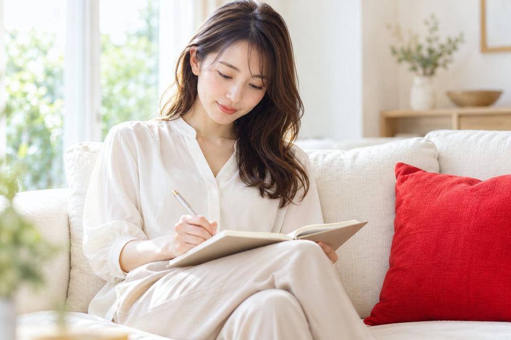ソファでノートを開き、ペンで自分の気持ちを書き出して整理している若い女性