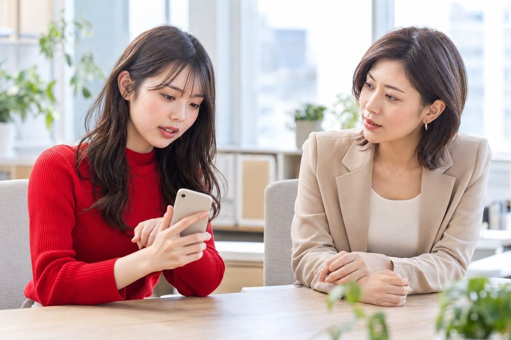 20代の女性がデスクでスマホを見ながら、落ち着いた表情でマネージャーに相談している