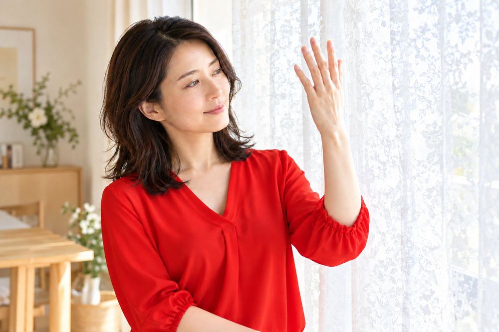 30代の女性が窓にレースのカーテンを引き、光がやわらかくなったか顔の前で手をかざして確かめている