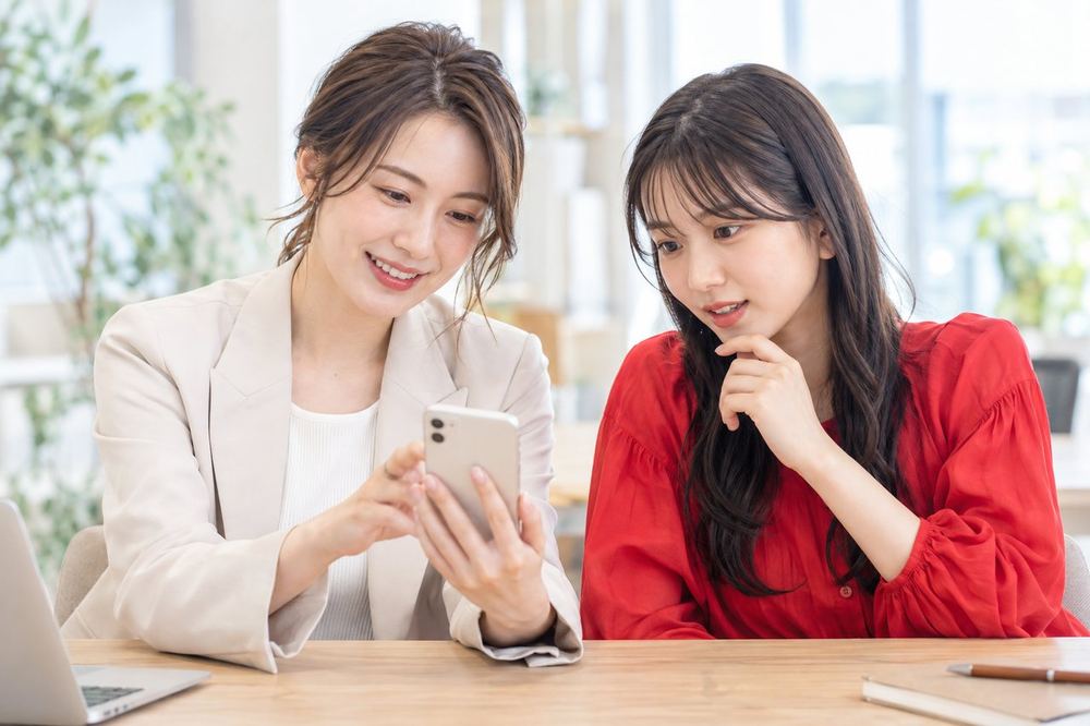 明るいオフィスで、ライバー経験のある女性のマネージャーが、20代の女性とスマホに映る夜の部屋の様子を見ながら話している
