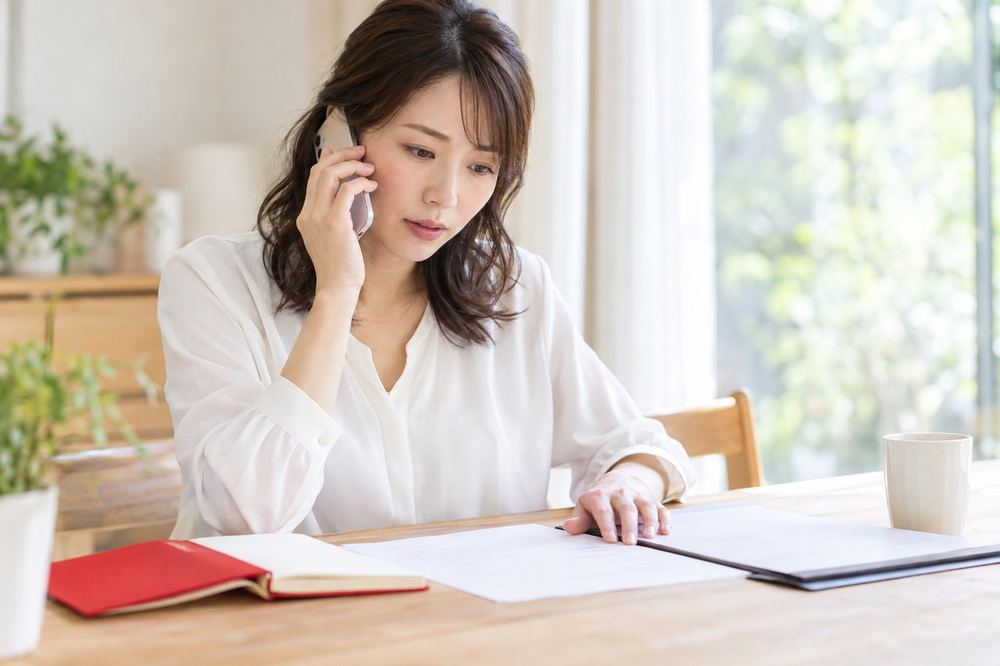 30代の女性が窓際でスマホを耳に当てて相談窓口に電話をしながら、手元のメモと契約書の控えを見ている