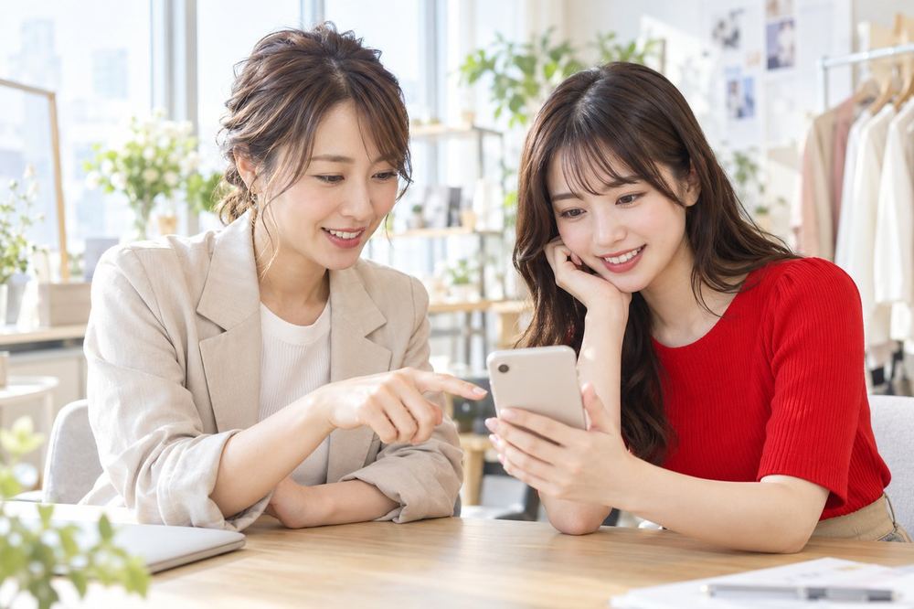 明るい部屋で、マネージャーの女性と新しく入る女性が、スマホを見ながら配信の準備を相談している