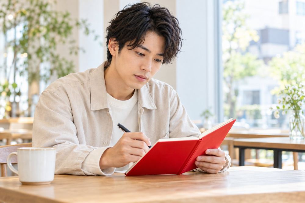 20代の男性がカフェの席で、事務所の面談で聞くことを書いたメモを見ながら質問を整理している
