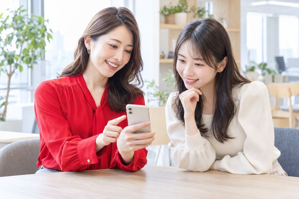 明るいオフィスで、ライバー経験のある女性のマネージャーが、20代の女性にスマホを見せながら報酬のしくみを説明している
