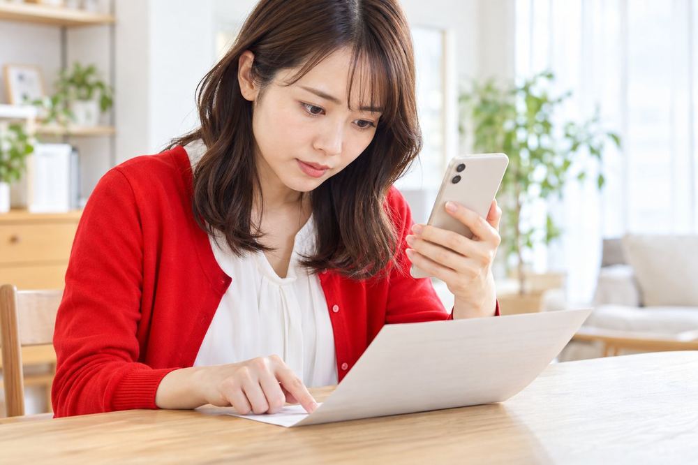 机で若い女性が、銀行口座の名義が書かれた書類を見ながら、スマホの登録内容と一字ずつ見比べている