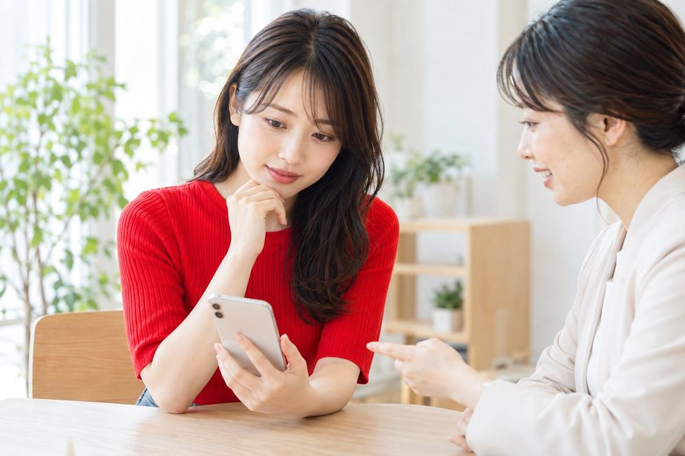 20代の女性が机でスマホを見ながら、落ち着いた表情で誰かと相談している