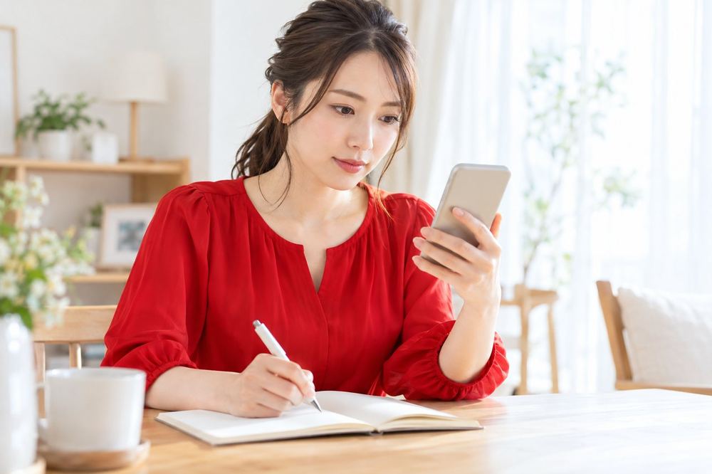 女性が自宅のテーブルで、スマホの通知画面を一つずつ確かめながらメモを取っている
