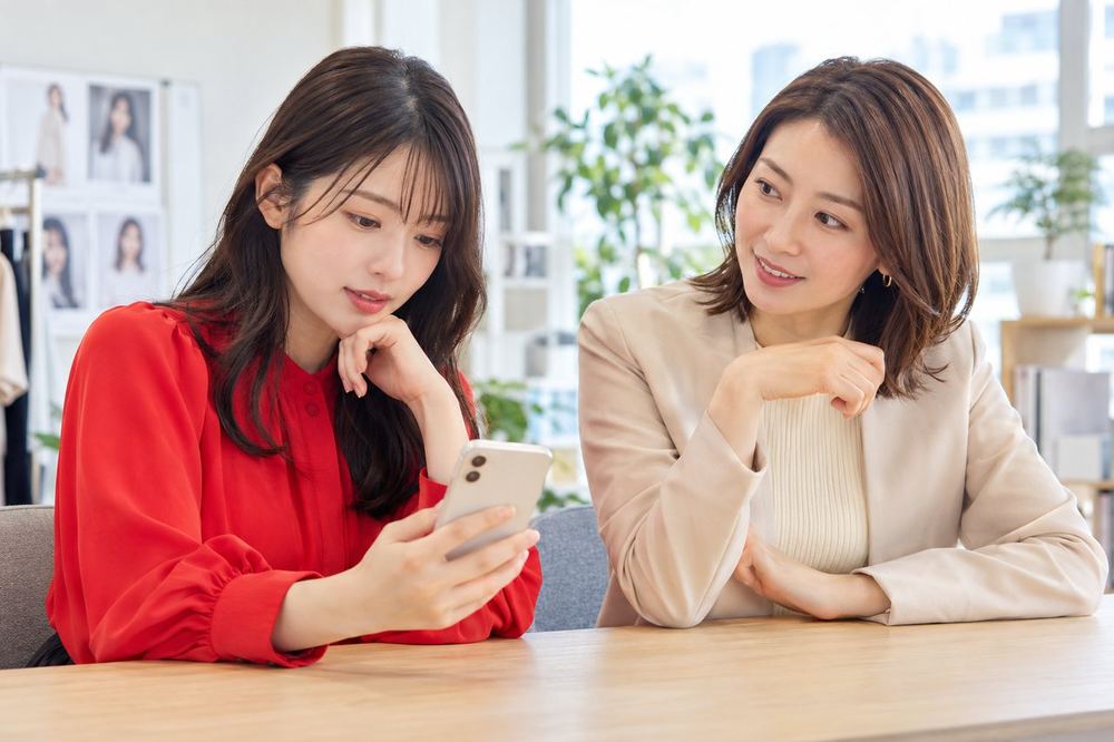 20代の女性がデスクでスマホを見ながら、落ち着いた表情でマネージャーと相談している