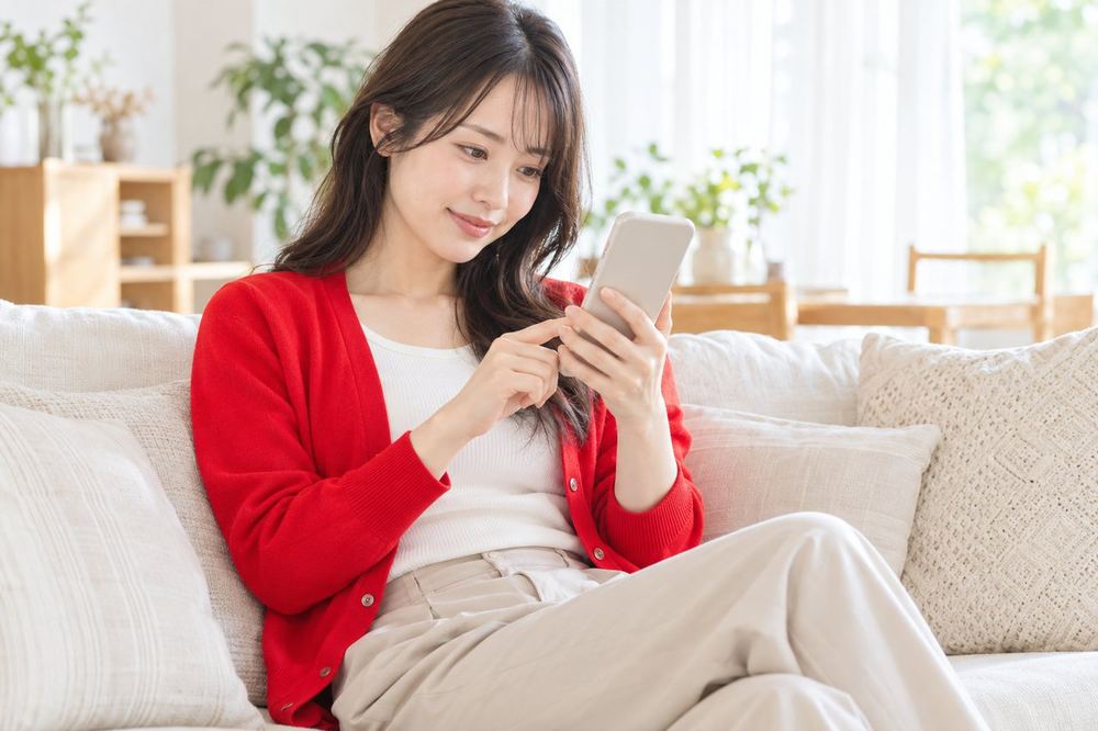 明るい部屋で女性がソファに座り、スマホの画面を見ながらコラボ配信に誘う相手を選んでいる