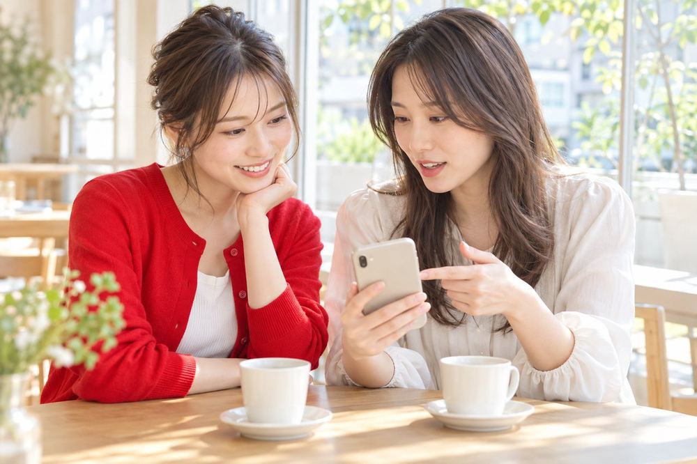 カフェの窓際で、ふたりの女性がスマホを見ながら、コラボ配信の日時と進め方を打ち合わせている