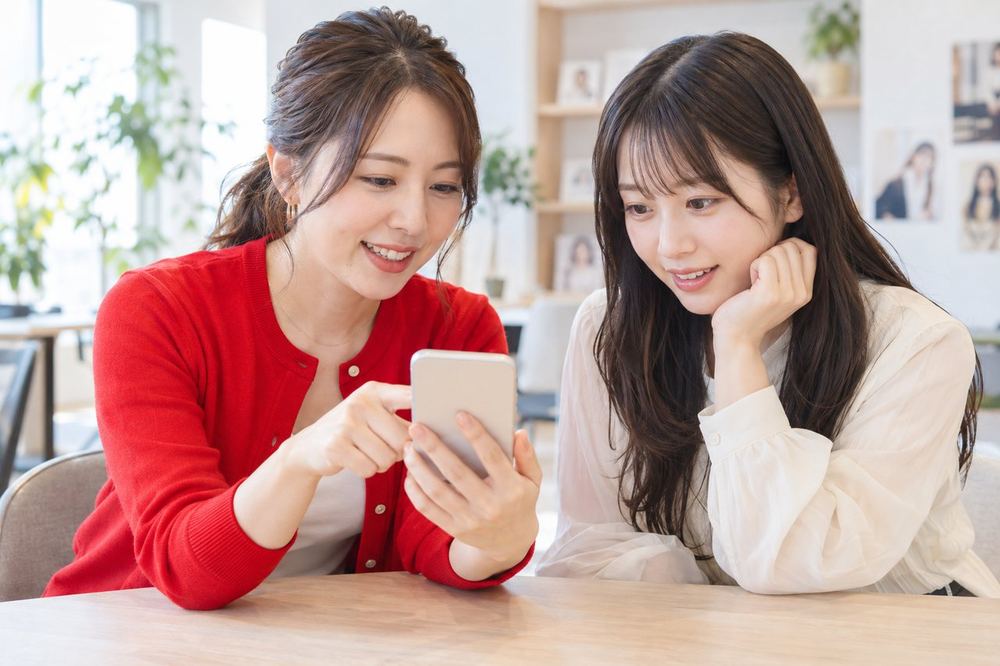 明るいオフィスで、ライバー経験のある女性のマネージャーが、20代の女性にスマホのLIVE画面を見せながらコメントのやりとりについて話している
