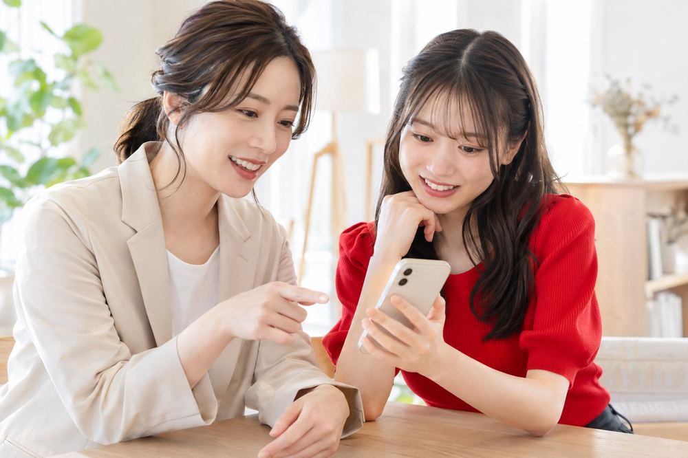 明るい部屋でマネージャーの女性と新人の女性がスマホを見ながら配信の準備を相談している