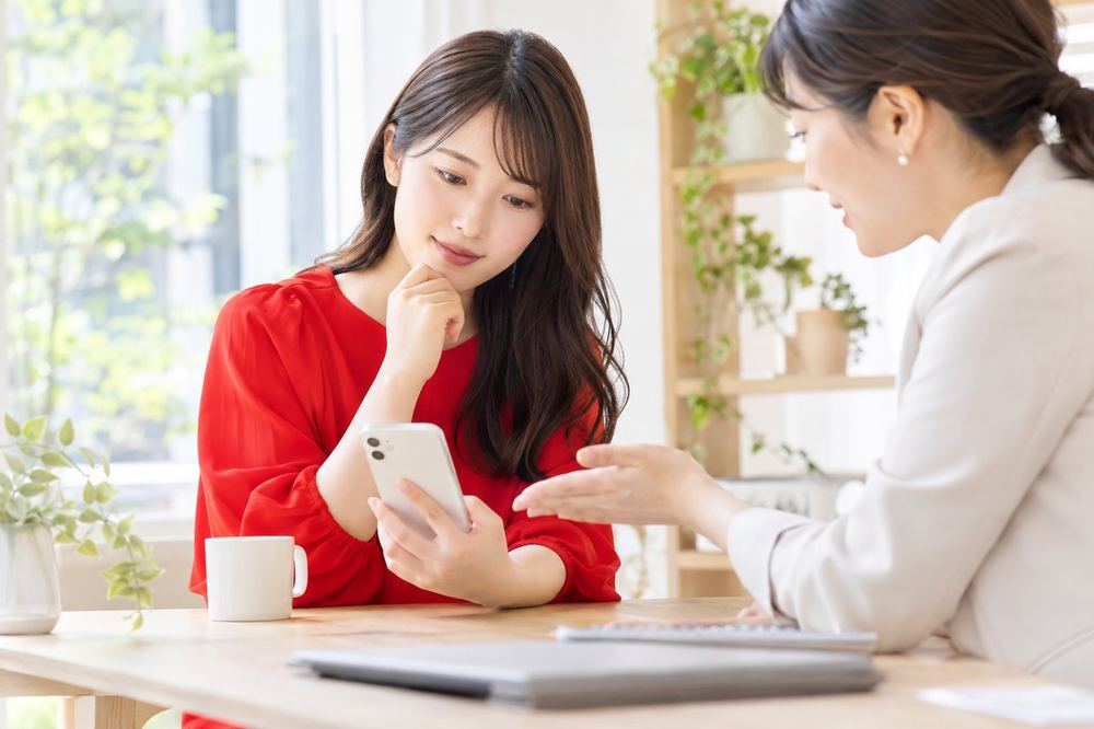 20代の女性がデスクでスマホを見ながら、落ち着いた表情で誰かと相談している