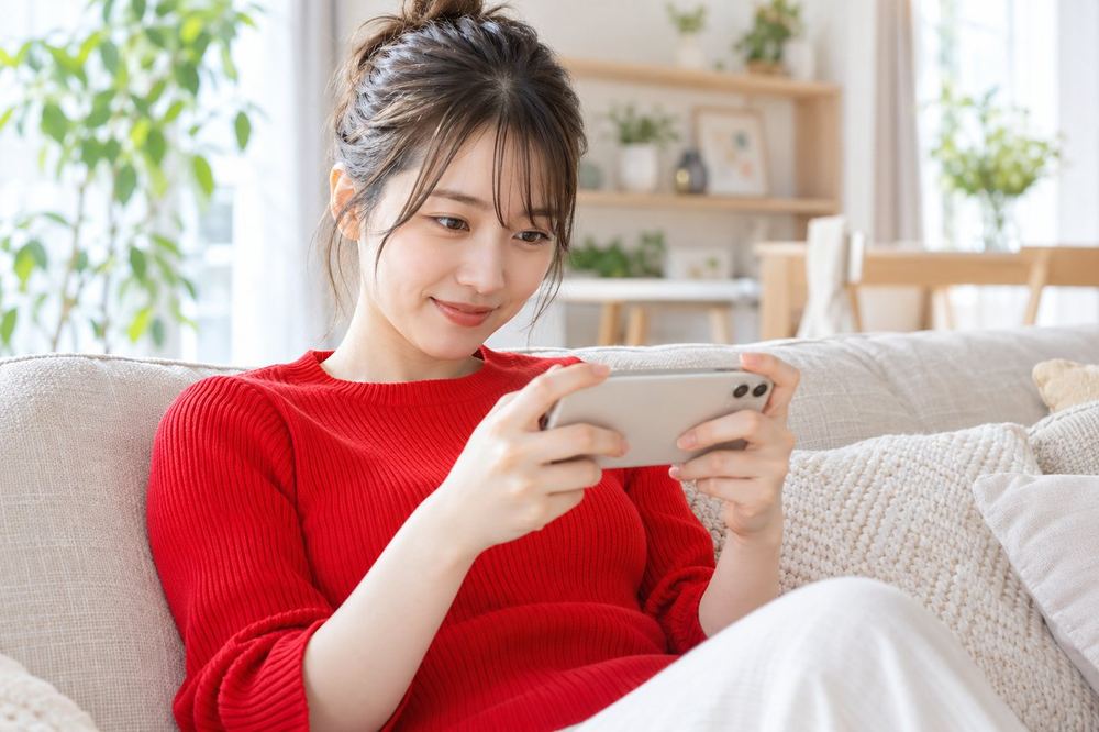20代の女性がソファでスマホを横向きに持ち、ゲームを遊びながら画面の通知を確認している