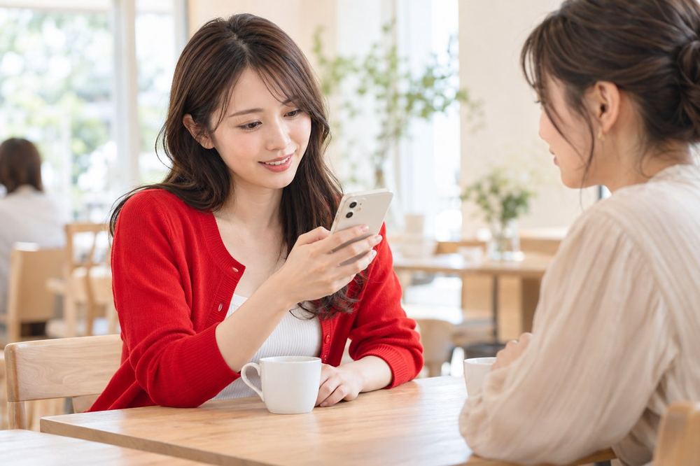 20代の女性がカフェでスマホを見ながら、向かいの人と配信の準備について話している