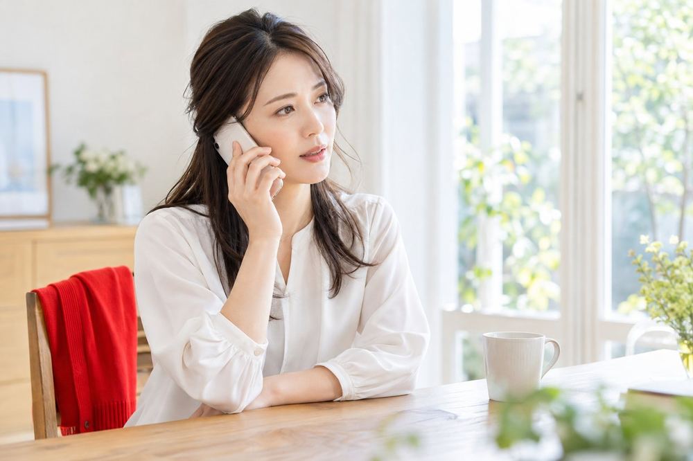 30代の女性が窓際の席でスマホを手に、落ち着いた表情で電話越しに相談している