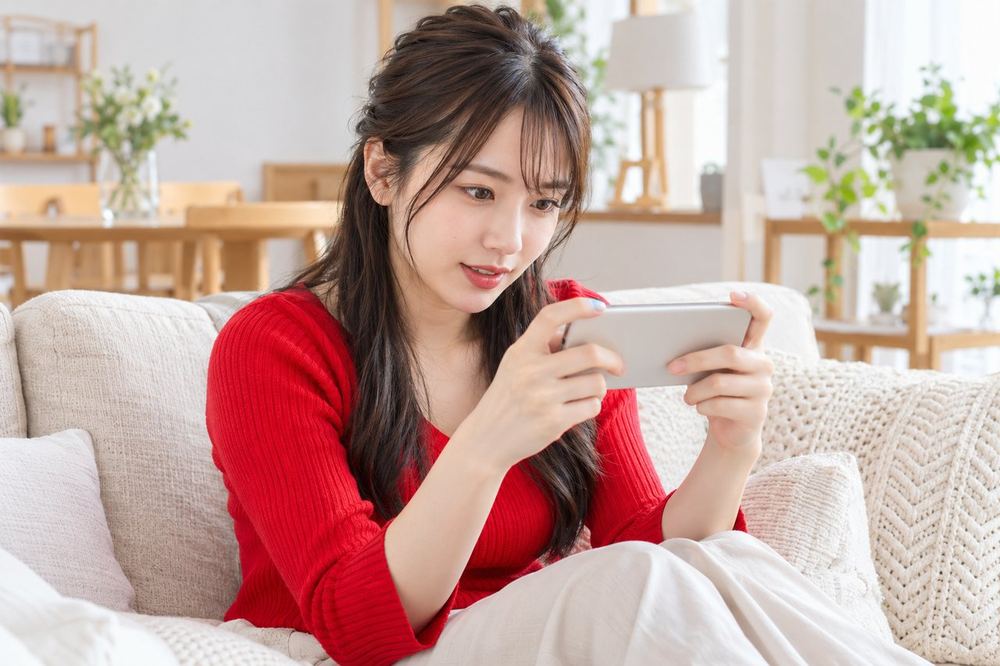 20代の女性が部屋のソファでスマホを横向きに持ち、バトル中のLIVE画面を見ながら画面の得点を目で追っている