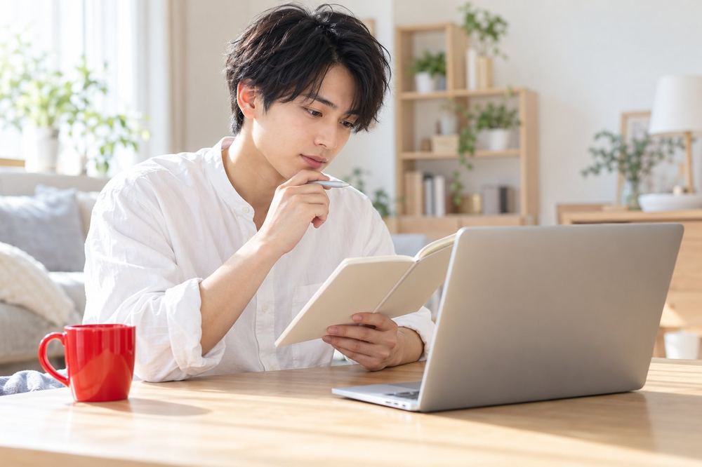 20代の男性が部屋でパソコンの前に座り、メモを見ながらバトルのしくみを整理している