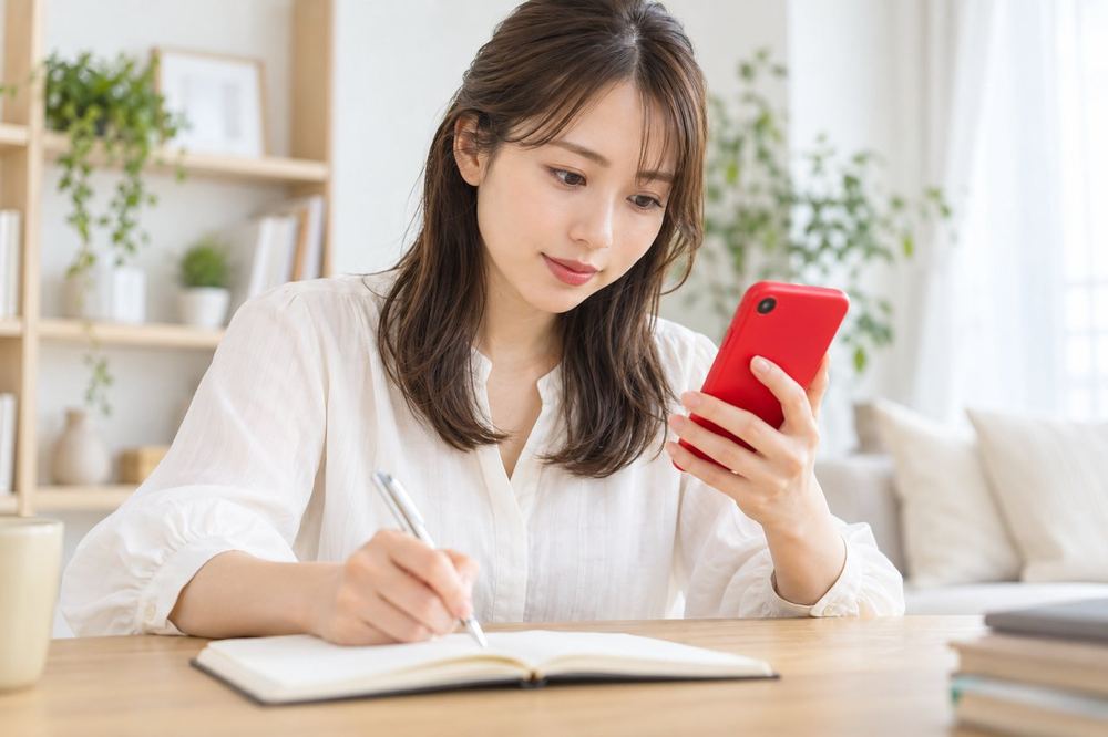 女性がスマホで複数の記事の数字を見比べながら、メモに出典を書き出している