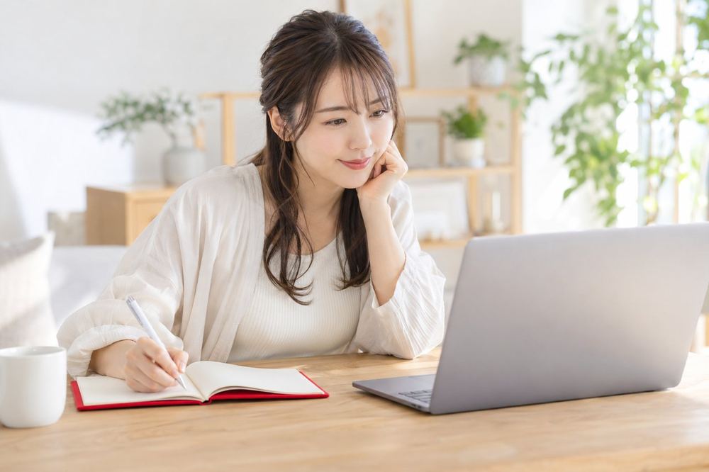 女性がノートパソコンの前で、前の配信の録画を見ながら、気づいたことをノートに書き留めている