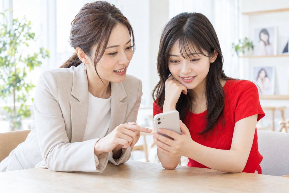 明るい部屋で、マネージャーの女性と新人の女性がスマホの配信画面を見ながら話し合っている