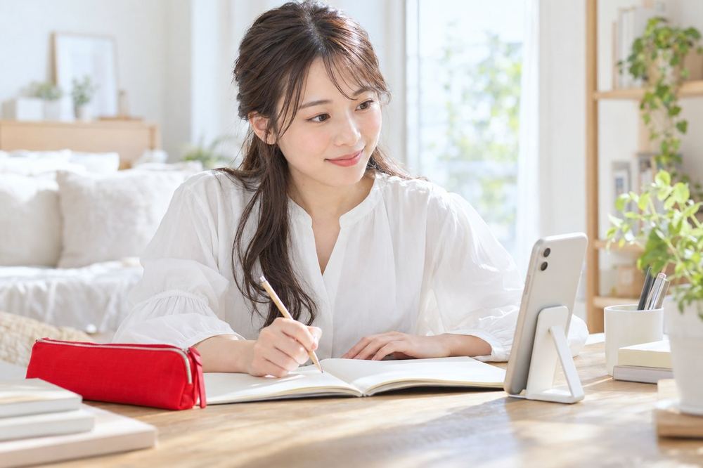 明るい部屋で女性が机に向かい、スマホを横に立てて、勉強しながらコメントを読んでいる